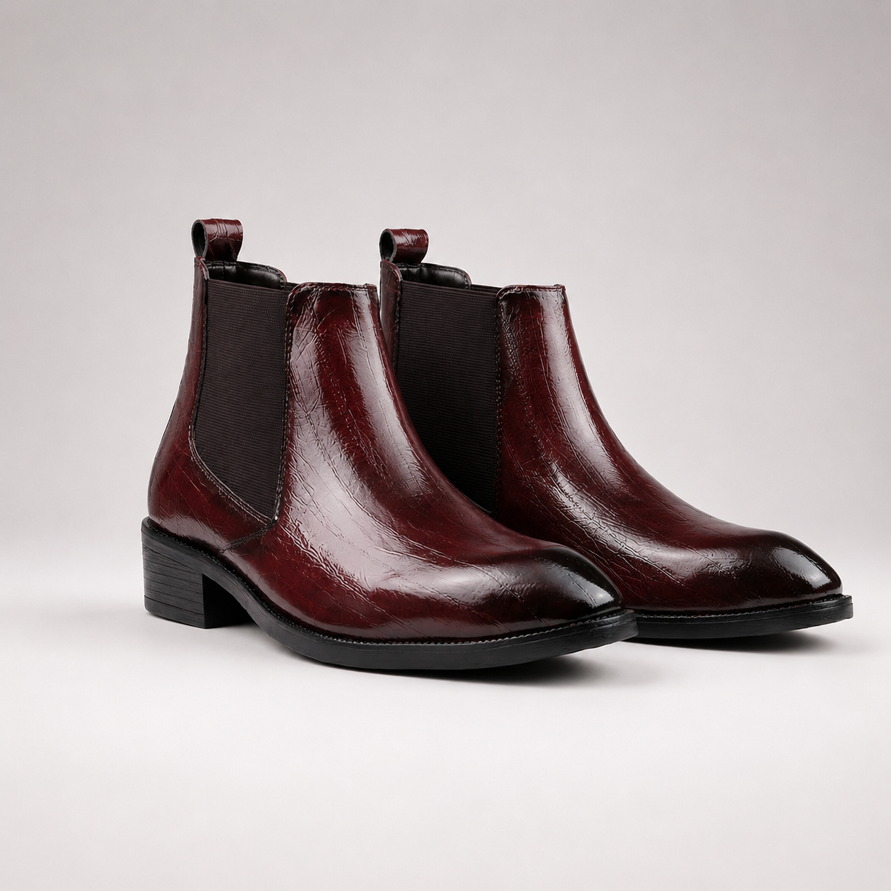 A pair of glossy dark burgundy leather Chelsea boots with black elastic side panels and low black block heels, photographed on a light gray studio background.