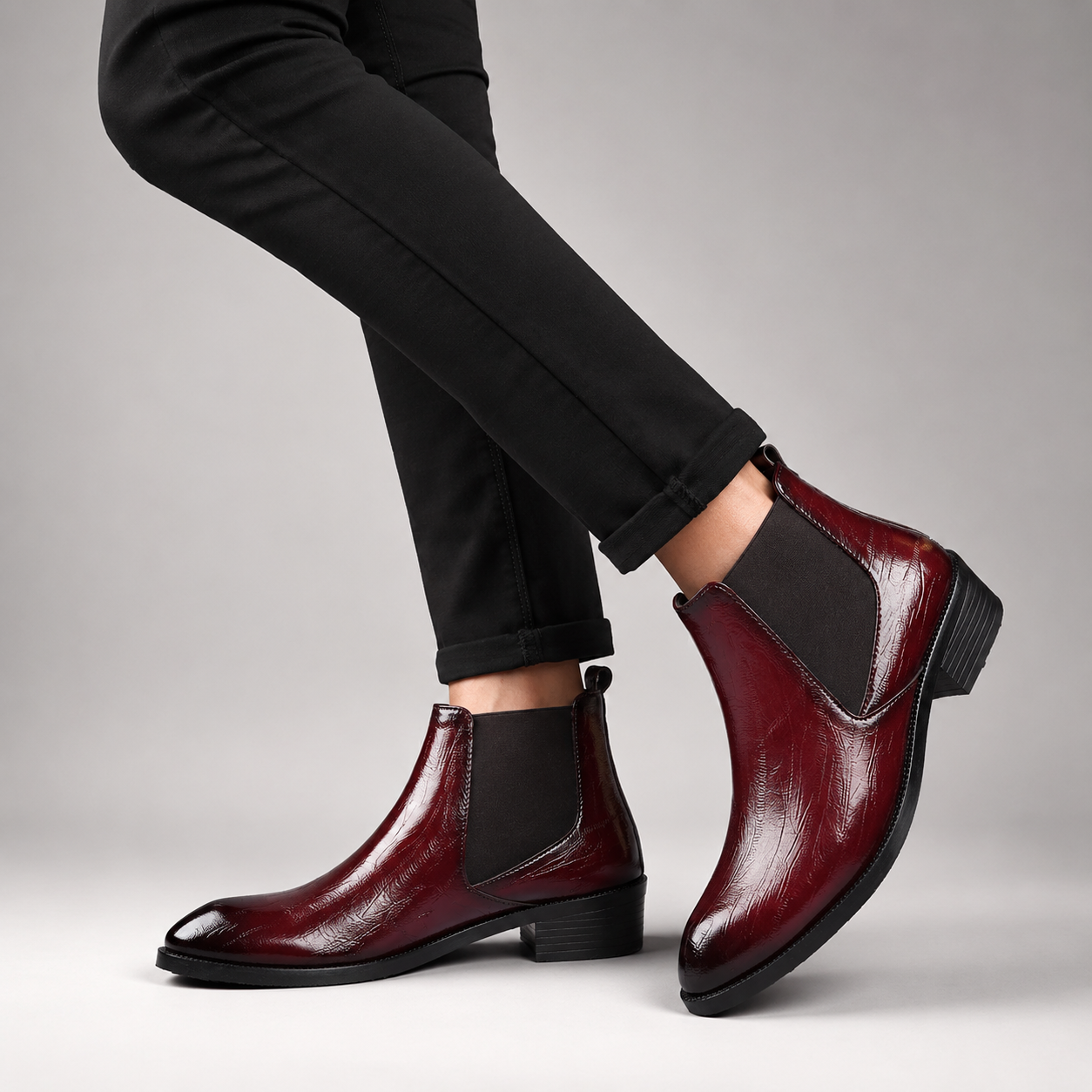 A pair of glossy burgundy Chelsea ankle boots with black elastic side panels, shown on a model wearing black pants against a neutral gray studio background.