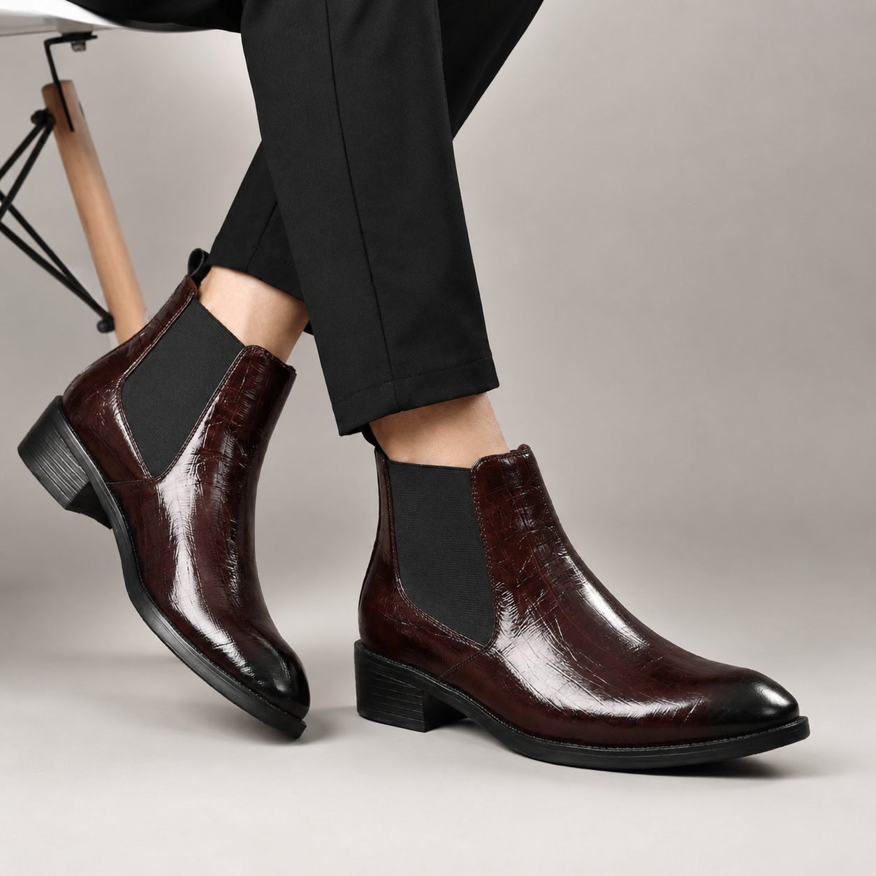 A pair of men's dark brown Chelsea boots with a glossy crocodile-embossed finish and black elastic side panels, shown on a model wearing black trousers against a neutral studio background.