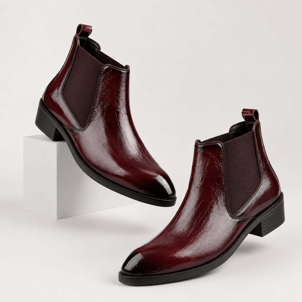 A pair of glossy dark burgundy leather Chelsea boots with black soles, displayed on a clean white studio background with one boot resting on a white block.