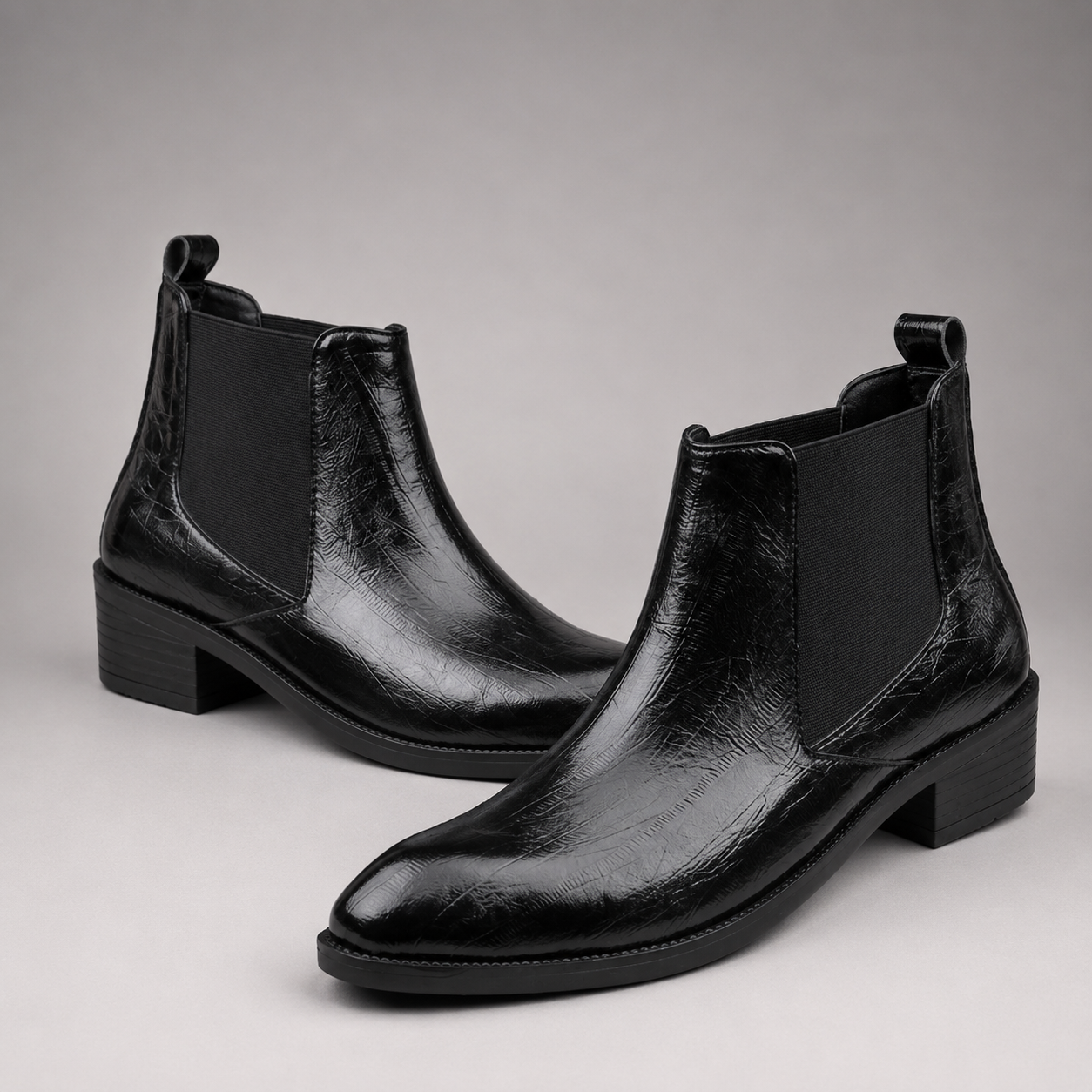 A pair of black leather Chelsea boots with a glossy, textured finish and black elastic side panels, shown on a neutral gray background.