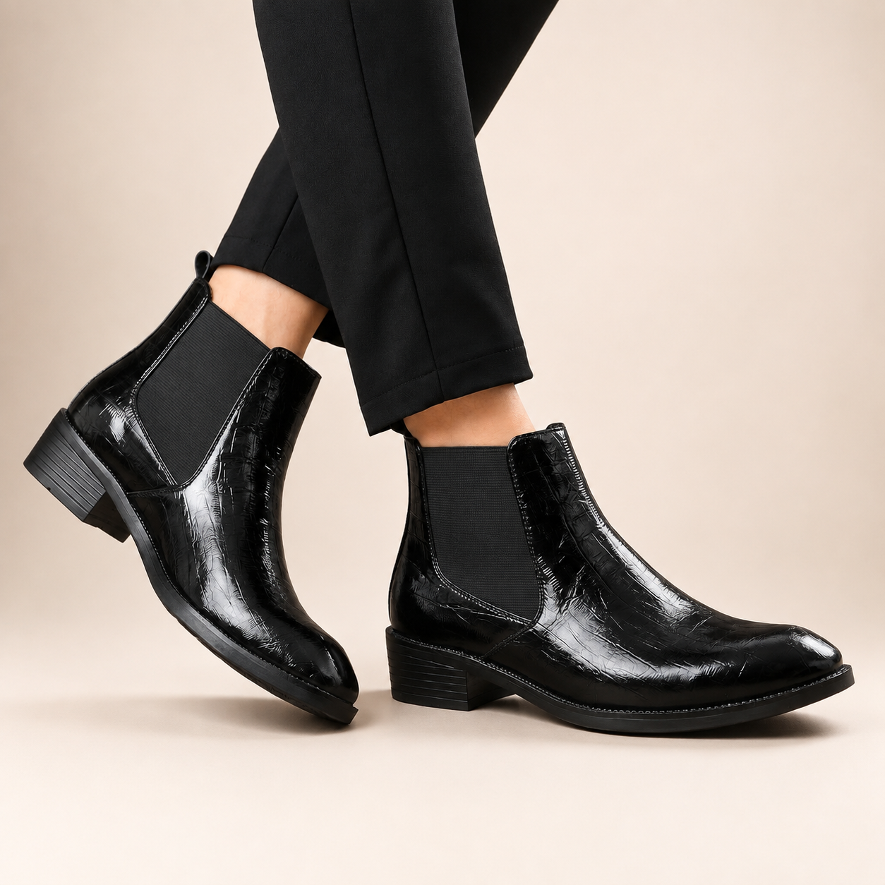 A pair of black glossy ankle boots with elastic side panels, shown on a model wearing black trousers against a warm beige studio background.