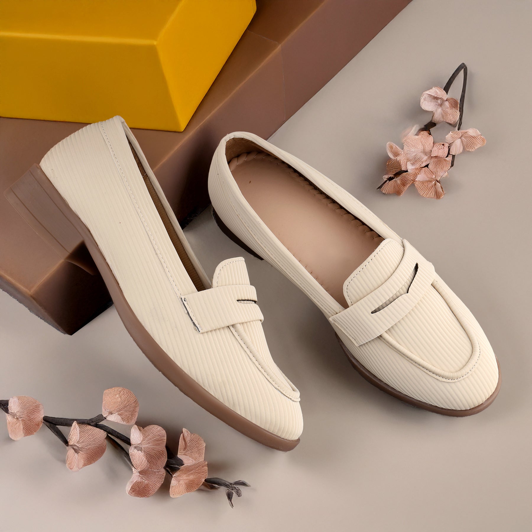 Pair of beige loafers on a light brown surface with a yellow box and floral decorations.