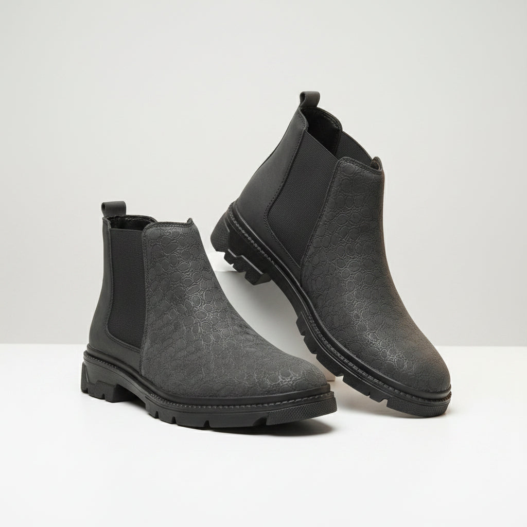A pair of dark gray textured Chelsea-style ankle boots with black elastic side panels and lug soles, displayed on a wooden surface against a soft beige curtain background.