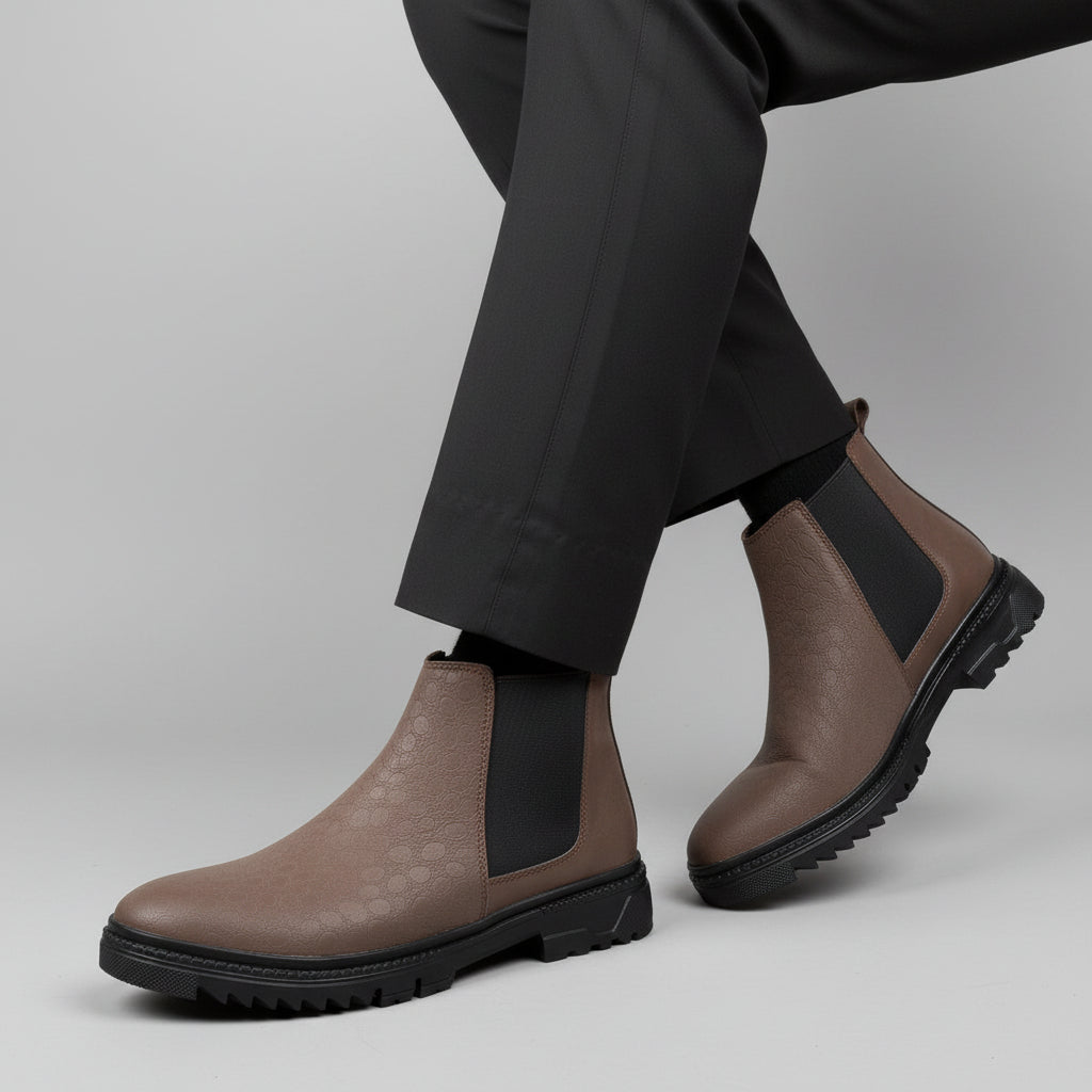 A person seated on a wooden chair wearing taupe-brown suede Chelsea-style ankle boots with black elastic side panels and black socks, photographed indoors on a wood floor against a textured white wall.