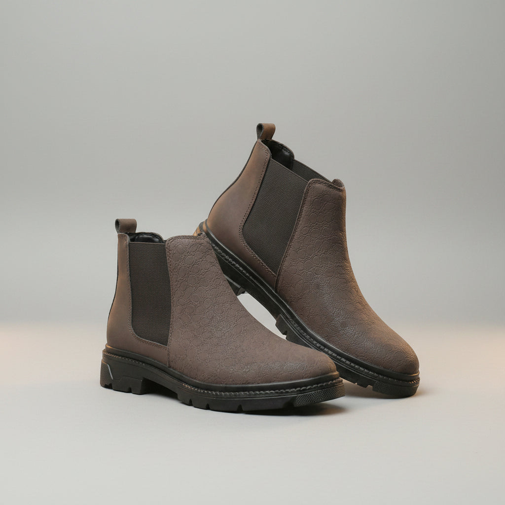 A pair of men's Chelsea-style ankle boots in taupe-brown textured leather with dark gray elastic side panels, displayed on a wooden surface against a soft beige curtain background.