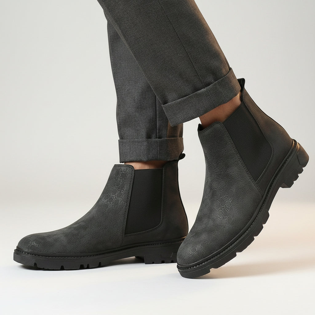 A pair of men's dark gray suede Chelsea boots with black elastic side panels, shown on a model wearing dark gray trousers on a wooden floor.
