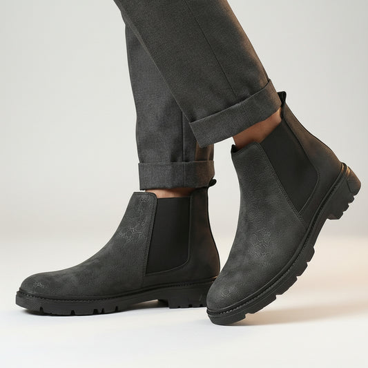 A pair of men's dark gray suede Chelsea boots with black elastic side panels, shown on a model wearing dark gray trousers on a wooden floor.