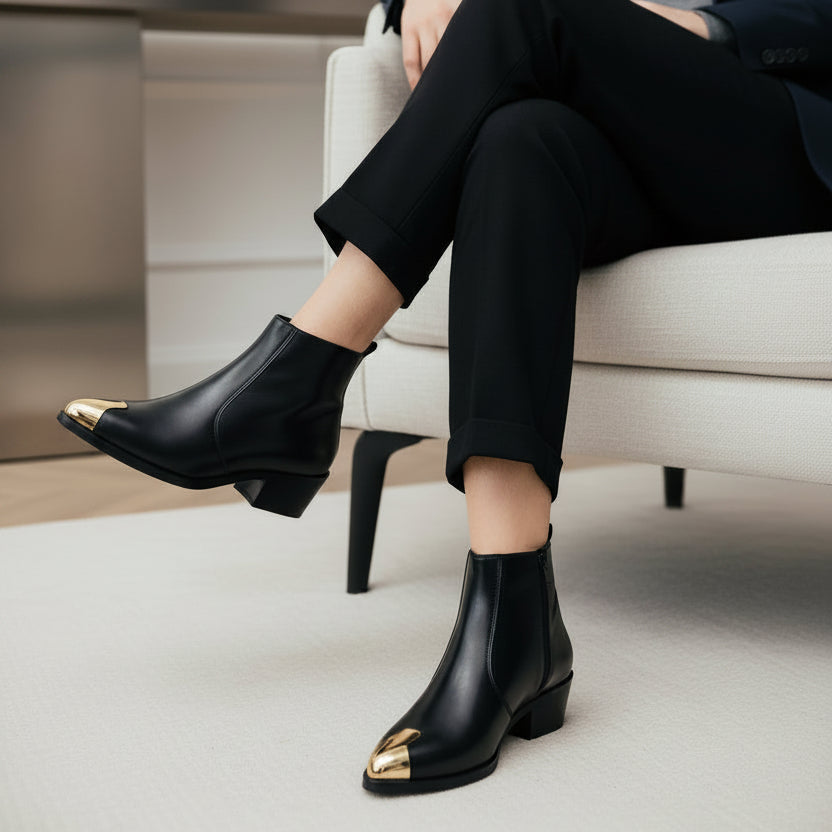 A pair of black leather ankle boots with metallic gold toe caps, with one boot being held by a hand and the other resting on a light wood floor. Yellow boxes are visible in the background.