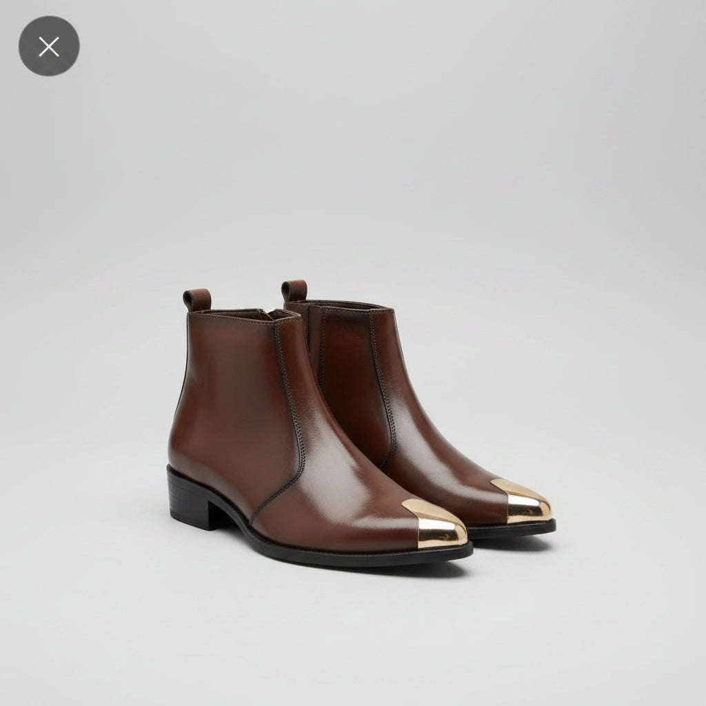 A pair of brown leather ankle boots with metallic gold toe caps, resting on a gray fabric sofa with a yellow box in the background.