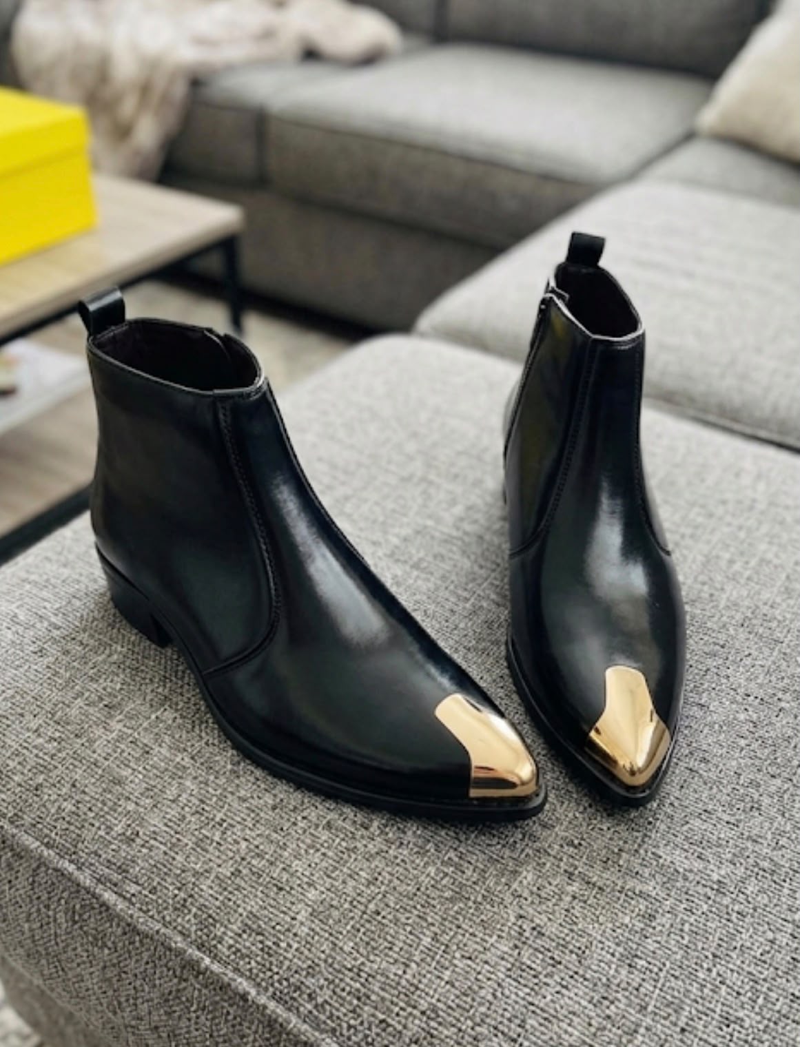 A pair of black leather ankle boots with metallic gold pointed toe caps, displayed on a gray fabric ottoman in a living room setting.