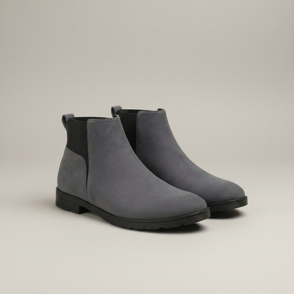 A pair of gray suede Chelsea-style ankle boots with black elastic side panels and black soles, displayed on a wooden surface with a neutral fabric runner against a beige background.