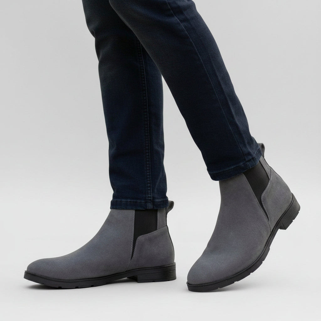 A person wearing dark gray suede Chelsea-style ankle boots with black elastic side panels, paired with dark blue jeans, standing on a cobblestone surface outdoors.