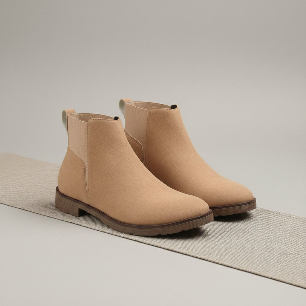 A pair of beige ankle boots with elastic side panels, displayed on a textured gray runner over a wooden surface against a warm beige background.