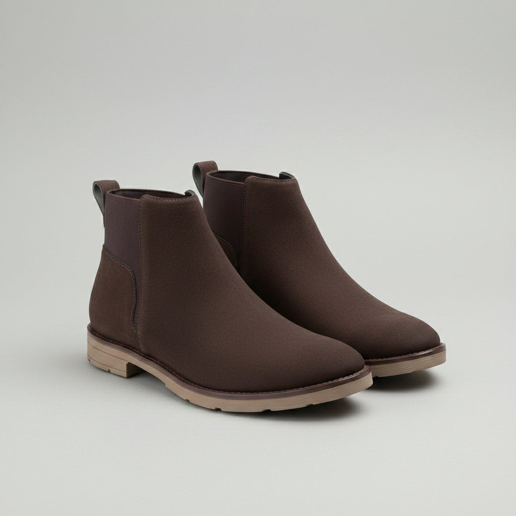A pair of dark brown suede Chelsea-style ankle boots with elastic side panels and low block heels, displayed on a wooden surface over a light textured fabric runner.