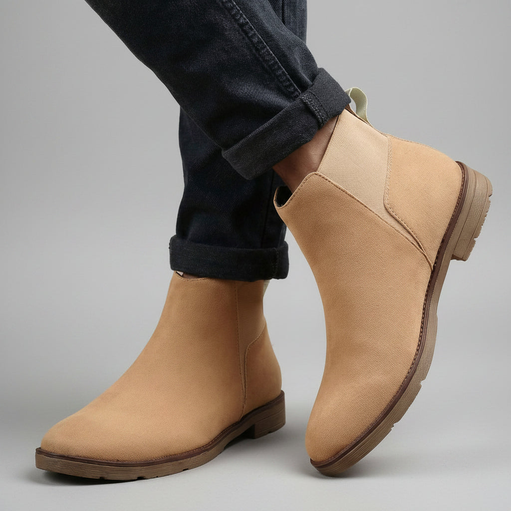 A pair of beige suede Chelsea-style ankle boots worn with dark cuffed jeans, shown against a neutral tan fabric backdrop.