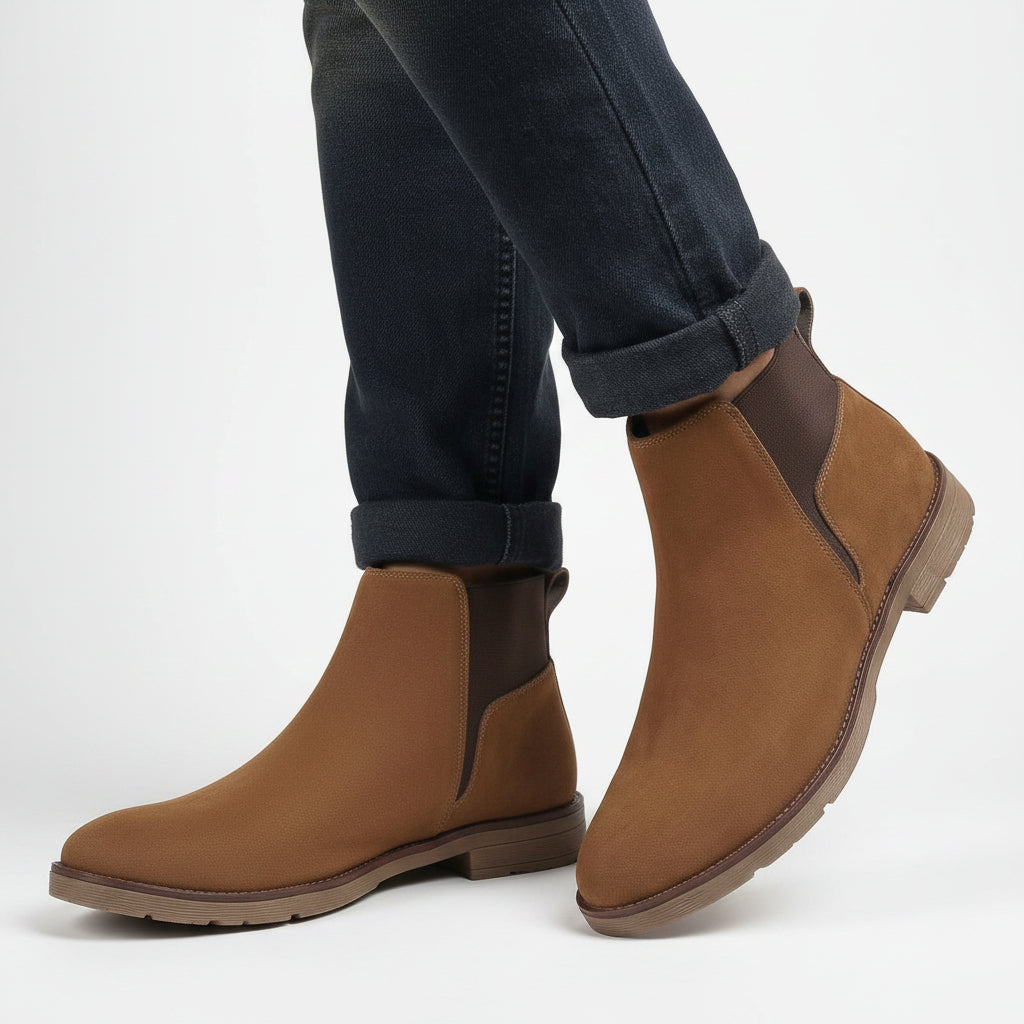 A person wearing brown suede Chelsea-style ankle boots with dark elastic side panels and dark blue cuffed jeans, standing against a warm beige fabric backdrop.
