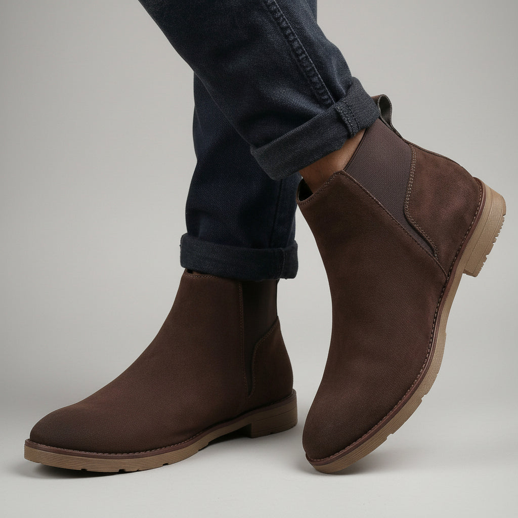 A pair of dark brown suede Chelsea-style ankle boots worn with dark cuffed jeans, set against a neutral beige fabric backdrop.