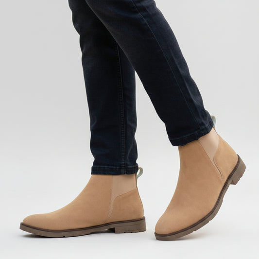 A person wearing tan suede Chelsea-style ankle boots with dark blue jeans, standing on a cobblestone path outdoors.