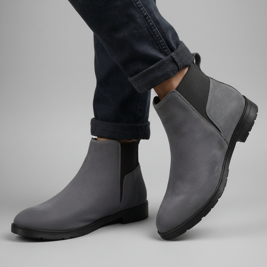 A pair of gray suede Chelsea-style ankle boots with black elastic side panels and black soles, shown on a model wearing dark cuffed jeans against a warm beige fabric backdrop.