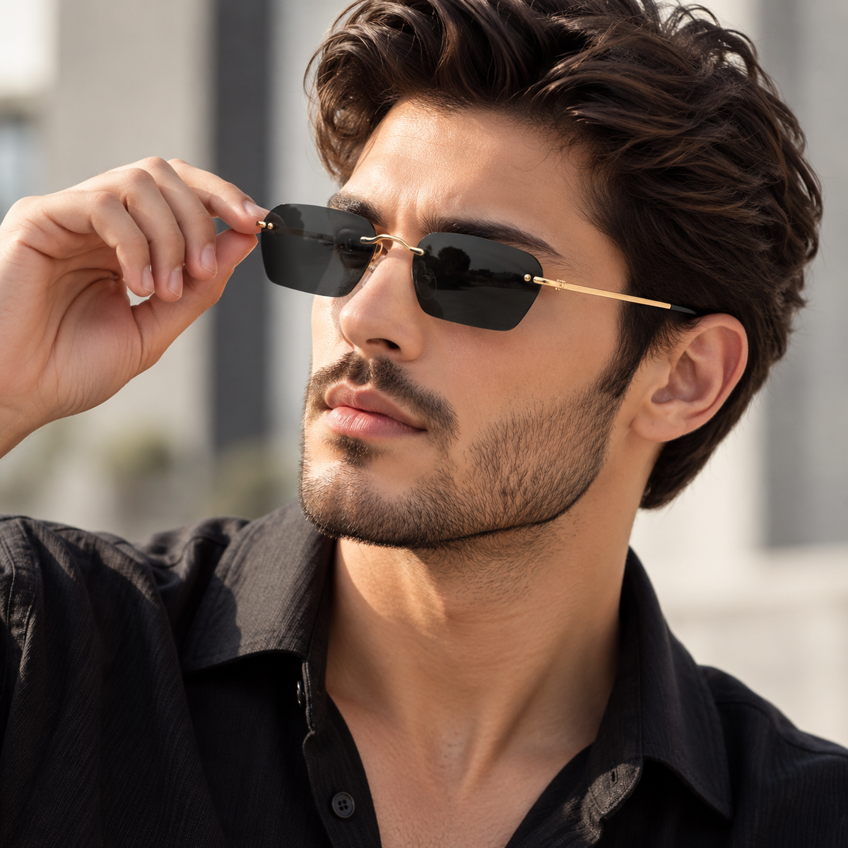 A man wearing rectangular black sunglasses with gold metal temples and rims, holding one lens with his hand. He is wearing a black button-down shirt against a blurred outdoor background.