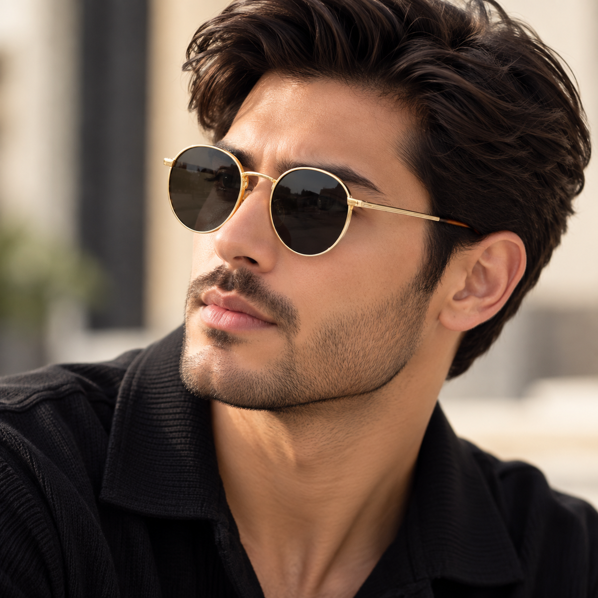 A close-up of a man wearing round gold-framed sunglasses with dark lenses and a black collared shirt, photographed outdoors with a softly blurred background.