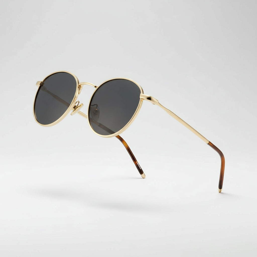 Oval sunglasses with gold metal frames, dark gray gradient lenses, and tortoiseshell temple arms, shown on a light gray background.