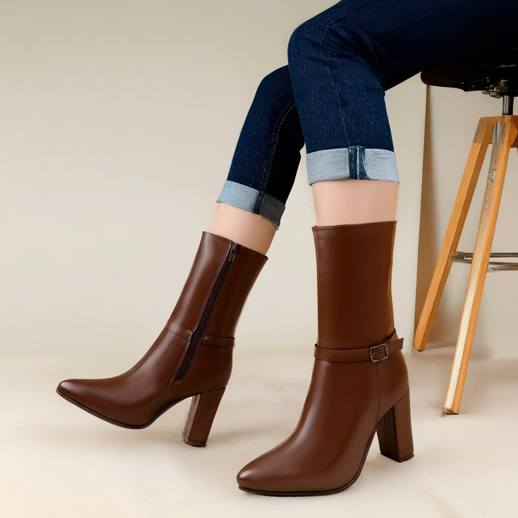 A pair of brown leather knee-high boots with block heels and side zippers, shown on a model wearing cuffed blue jeans against a neutral studio background.