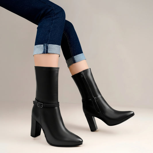 A pair of black leather mid-calf heeled boots with side buckle straps, shown on a model wearing cuffed blue jeans, seated on a wooden stool.