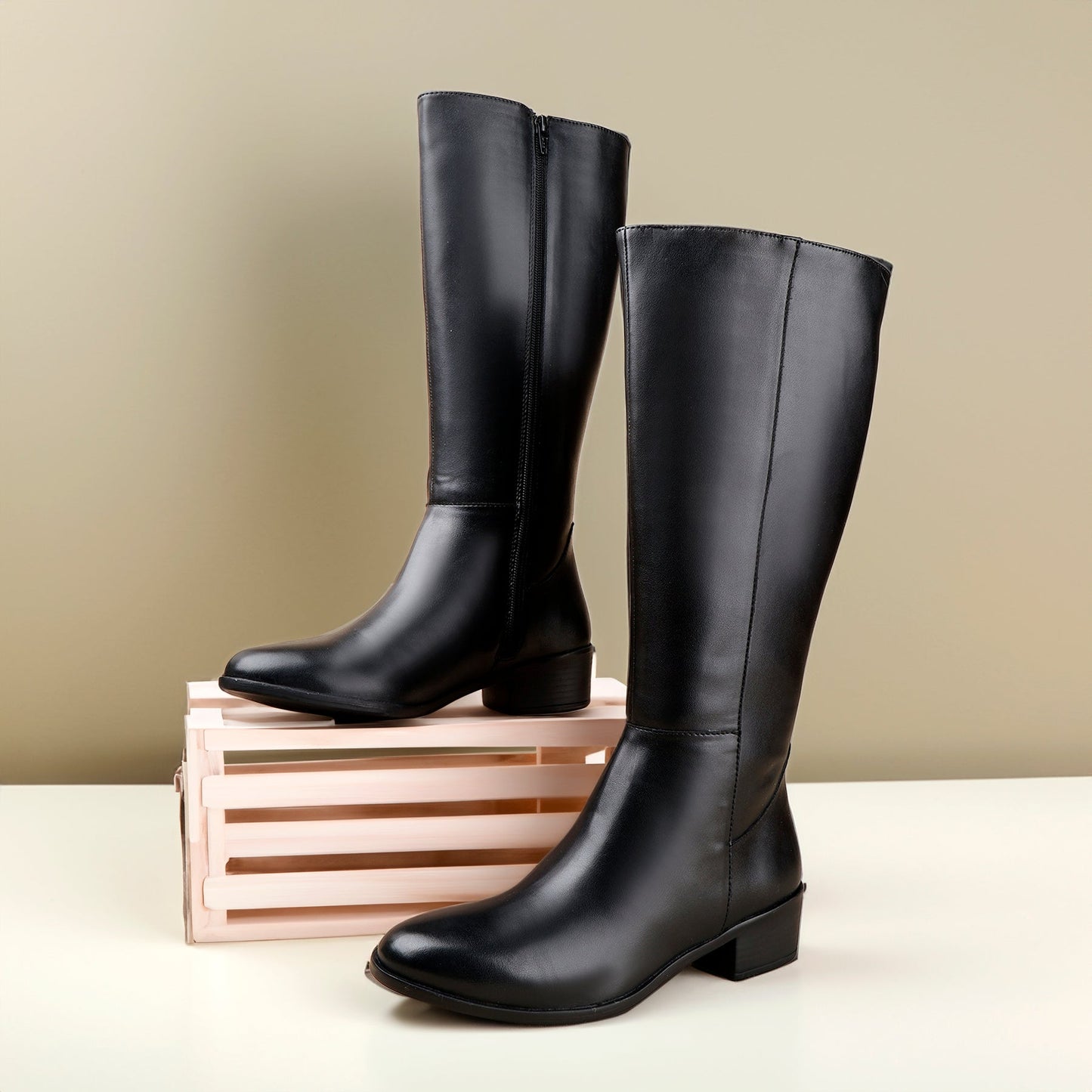 A pair of black leather knee-high boots with low block heels, displayed on a light pink wooden crate against a neutral beige background.