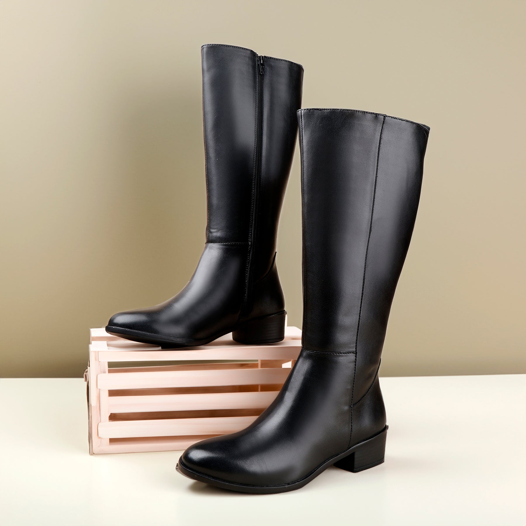 A pair of black leather knee-high boots with low block heels, displayed on a light pink wooden crate against a neutral beige background.
