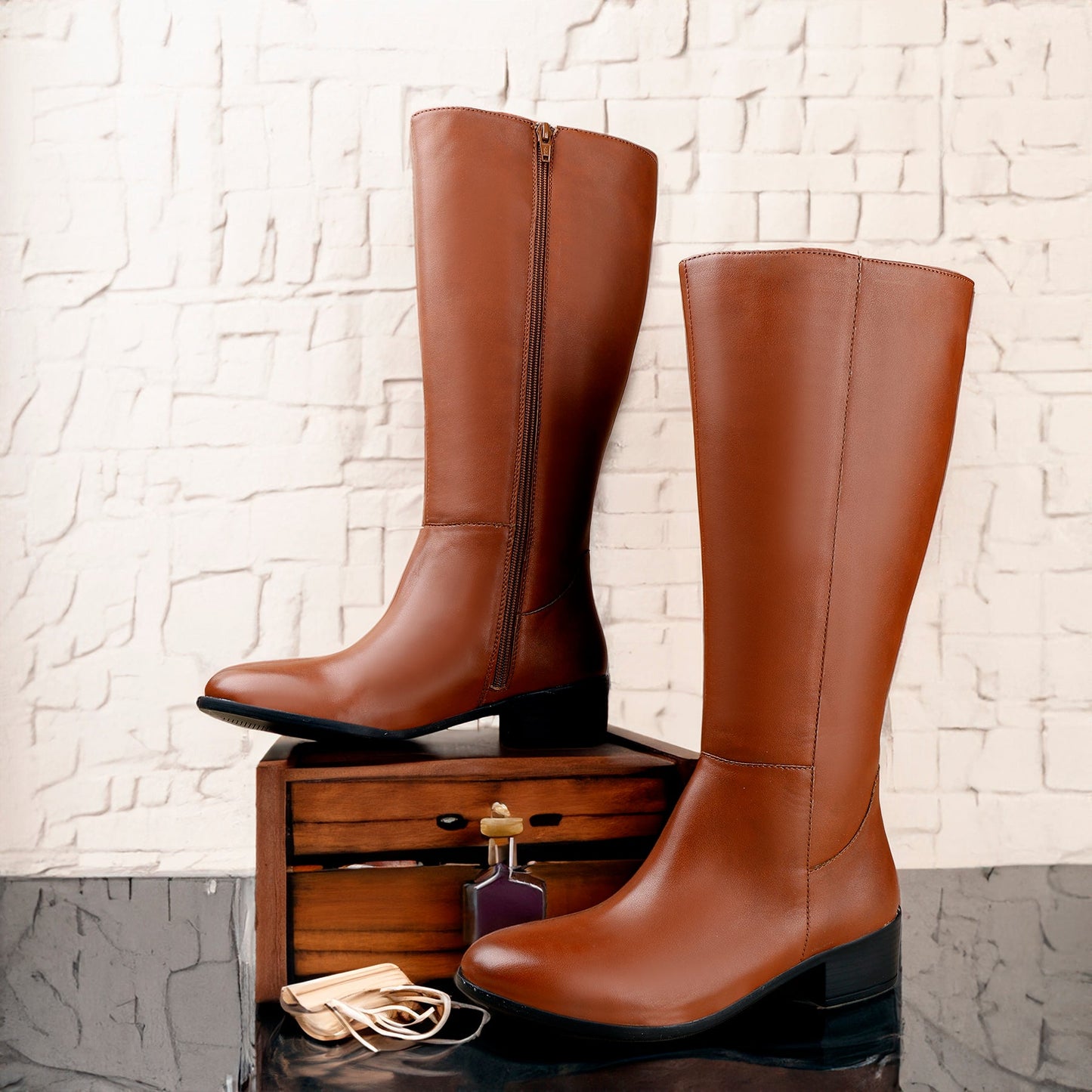 A pair of knee-high brown leather boots with side zippers, displayed on a small wooden box against a white textured brick wall.