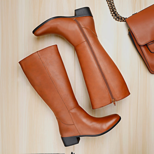 A pair of knee-high brown leather boots with side zippers and low block heels, arranged on a light wood background next to a matching brown handbag.