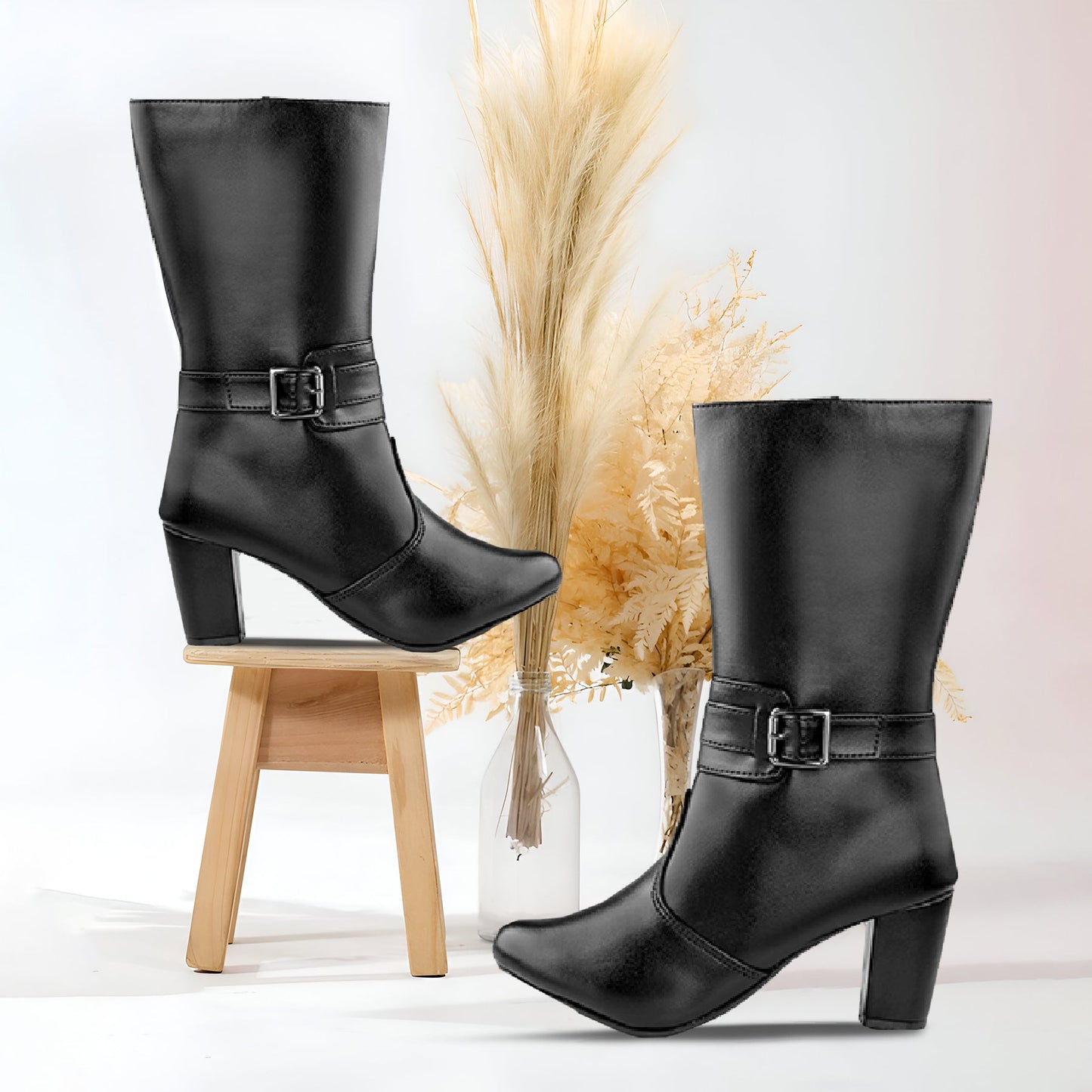 A pair of black knee-high leather boots with a block heel and a decorative side strap with a buckle, styled on a small wooden stool and a glass vase with dried pampas grass against a white background.