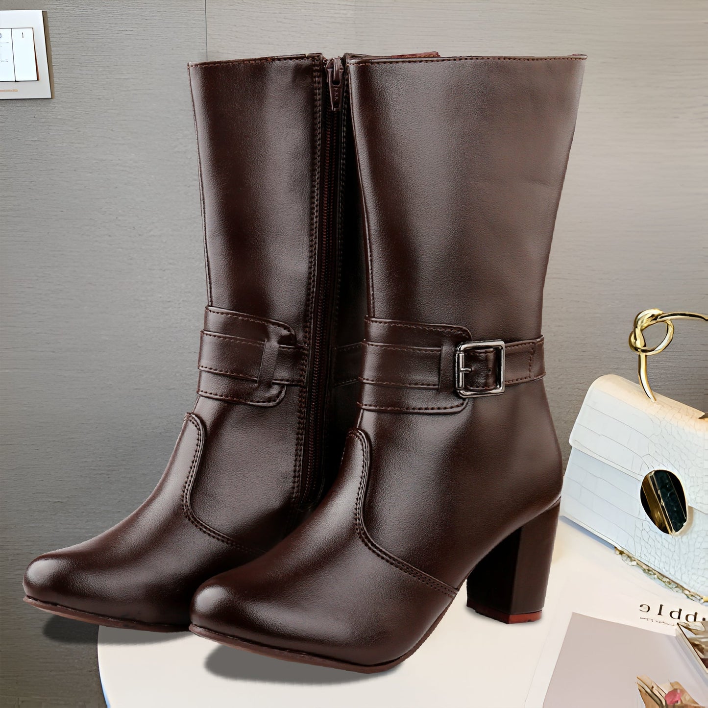 A pair of dark brown faux-leather mid-calf boots with block heels and side buckle straps, displayed on a white surface against a gray wall.