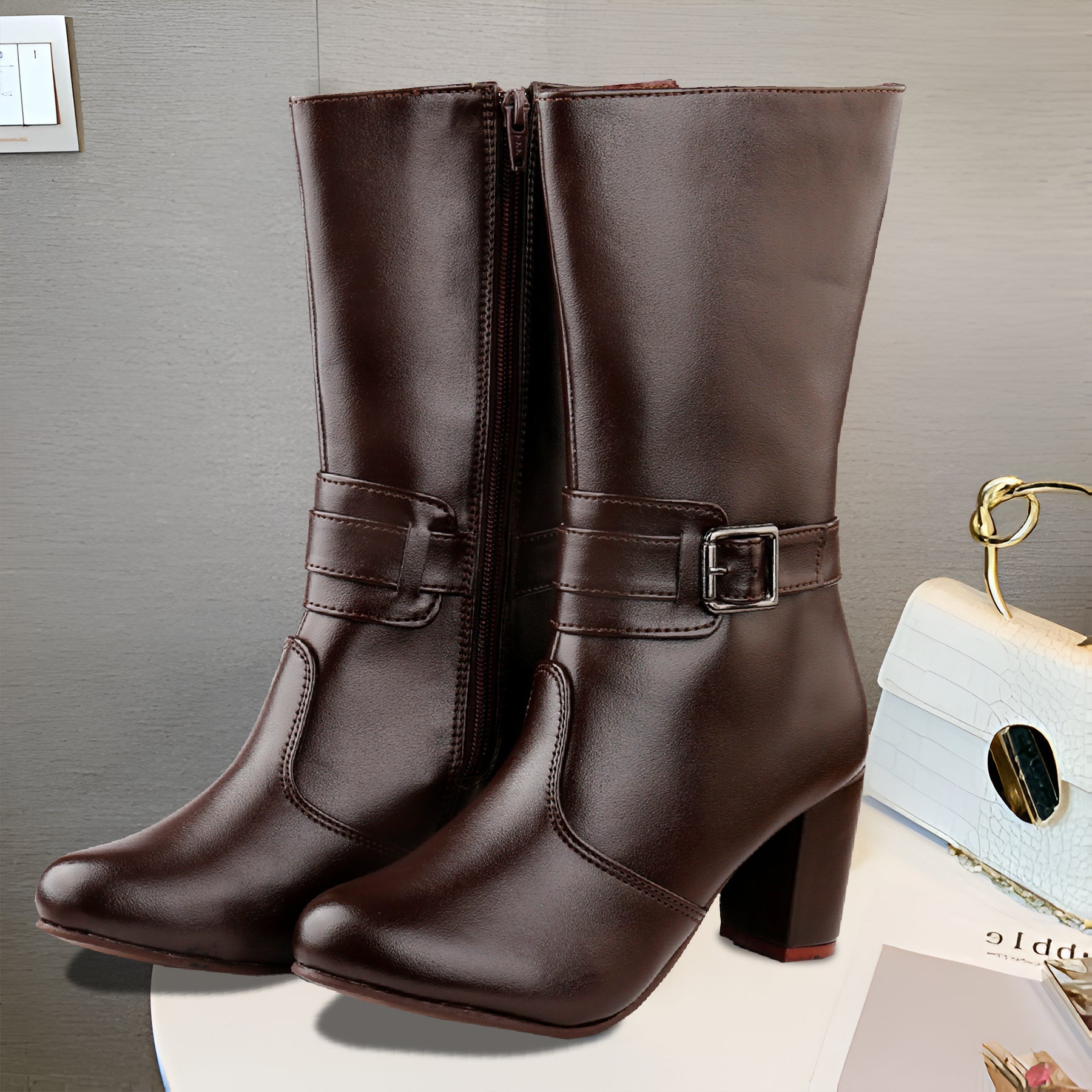 A pair of dark brown faux-leather mid-calf boots with block heels and side buckle straps, displayed on a white surface against a gray wall.