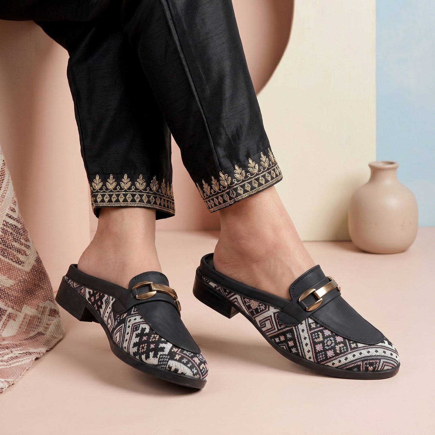 Black patterned clogs with gold accents worn with black pants against a neutral background