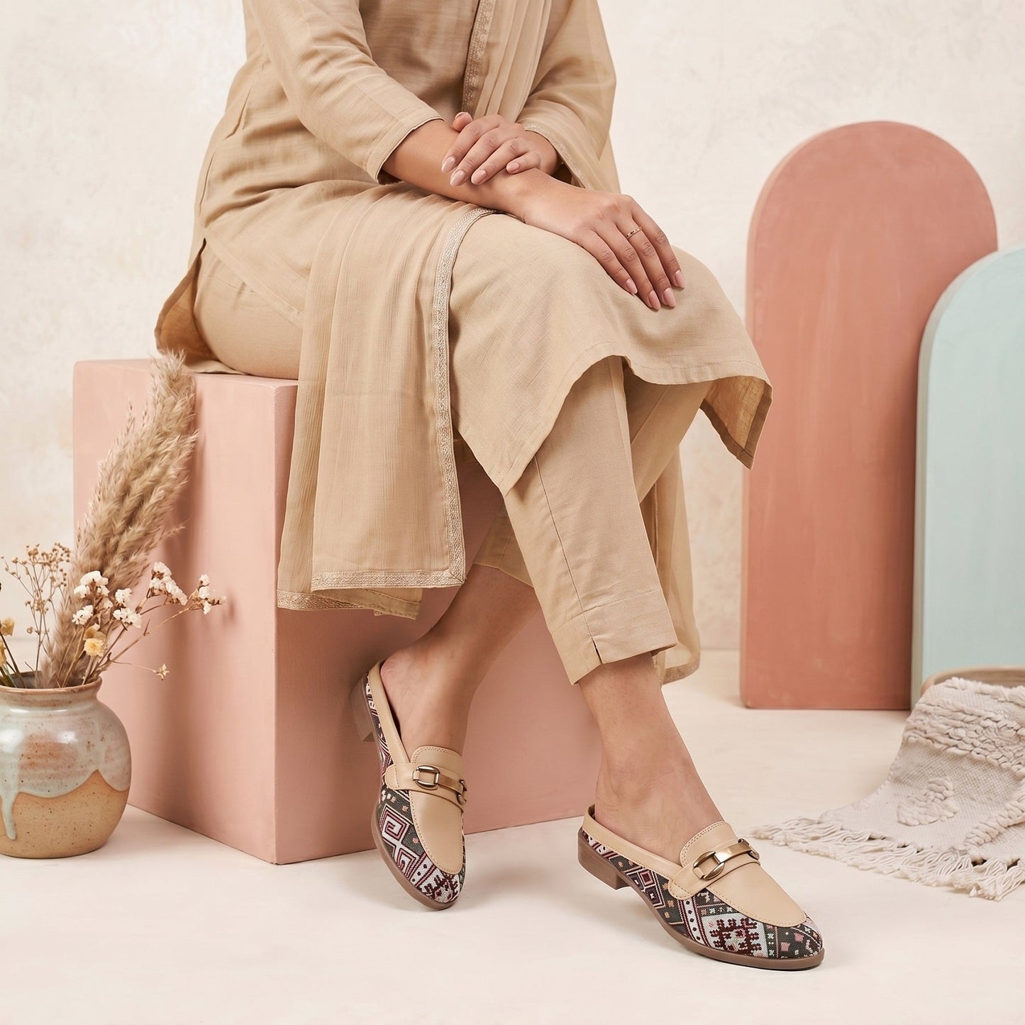 Person wearing a beige outfit with patterned shoes sitting on a pink block.