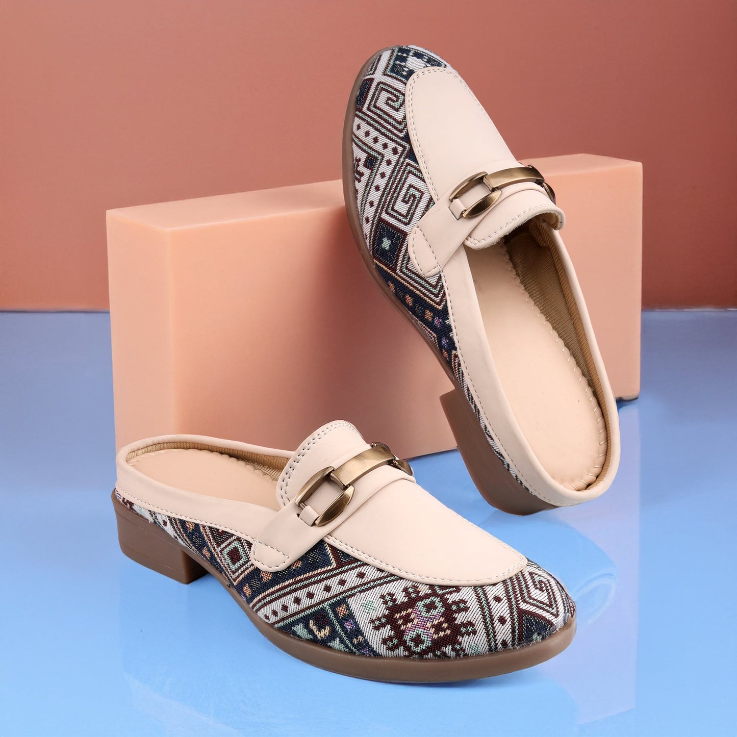 A pair of women's slip-on mule loafers with cream-colored leather uppers and a geometric patterned fabric lower panel, displayed on a blue reflective surface with a peach-colored block prop.