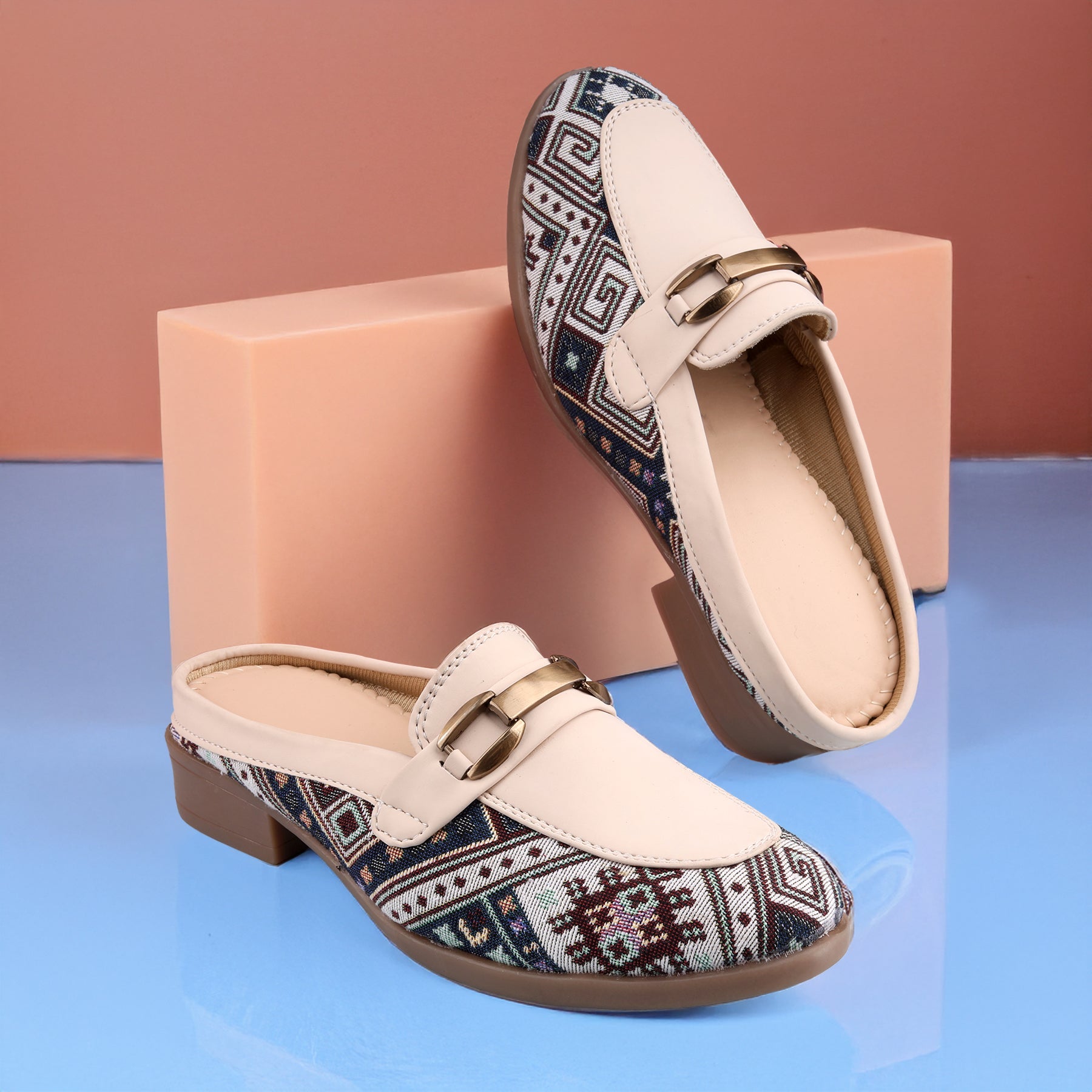 A pair of women's slip-on mule loafers with cream-colored leather uppers and a geometric patterned fabric lower panel, displayed on a blue reflective surface with a peach-colored block prop.