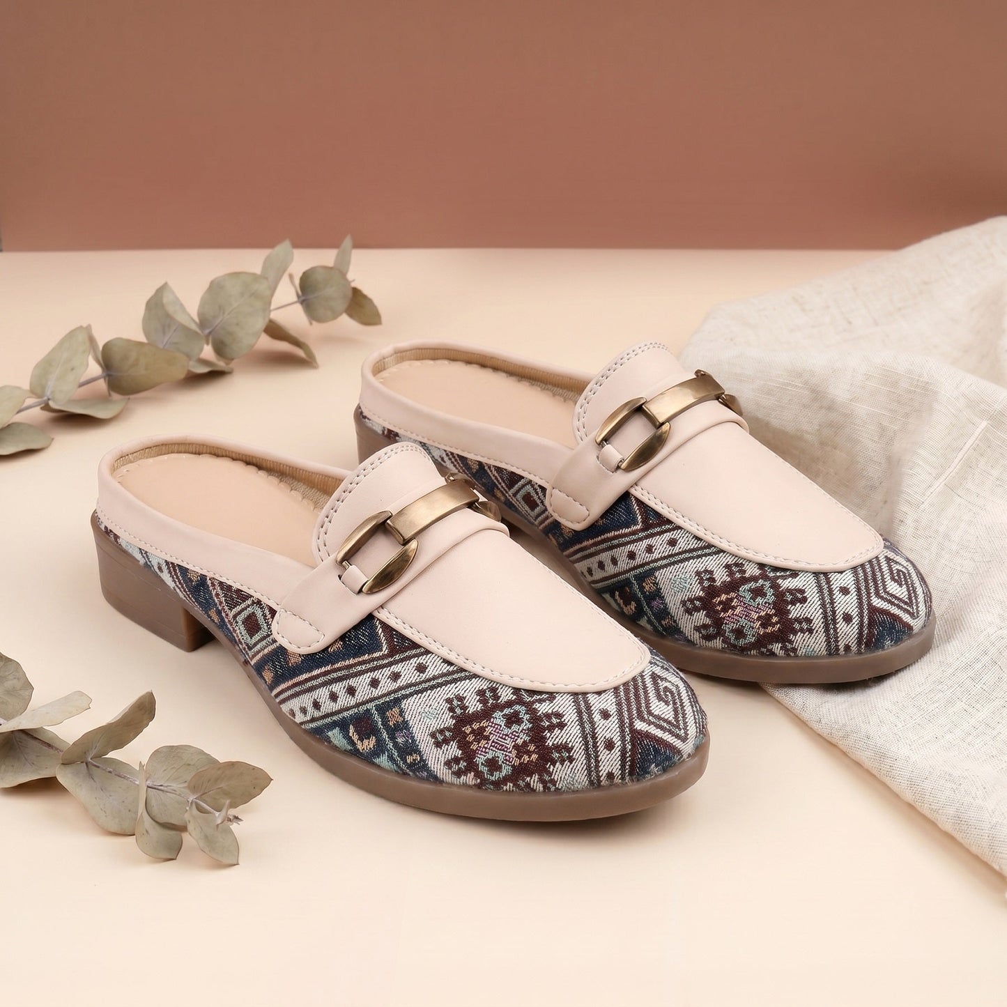 Pair of loafers with patterned soles on a beige surface with eucalyptus leaves.