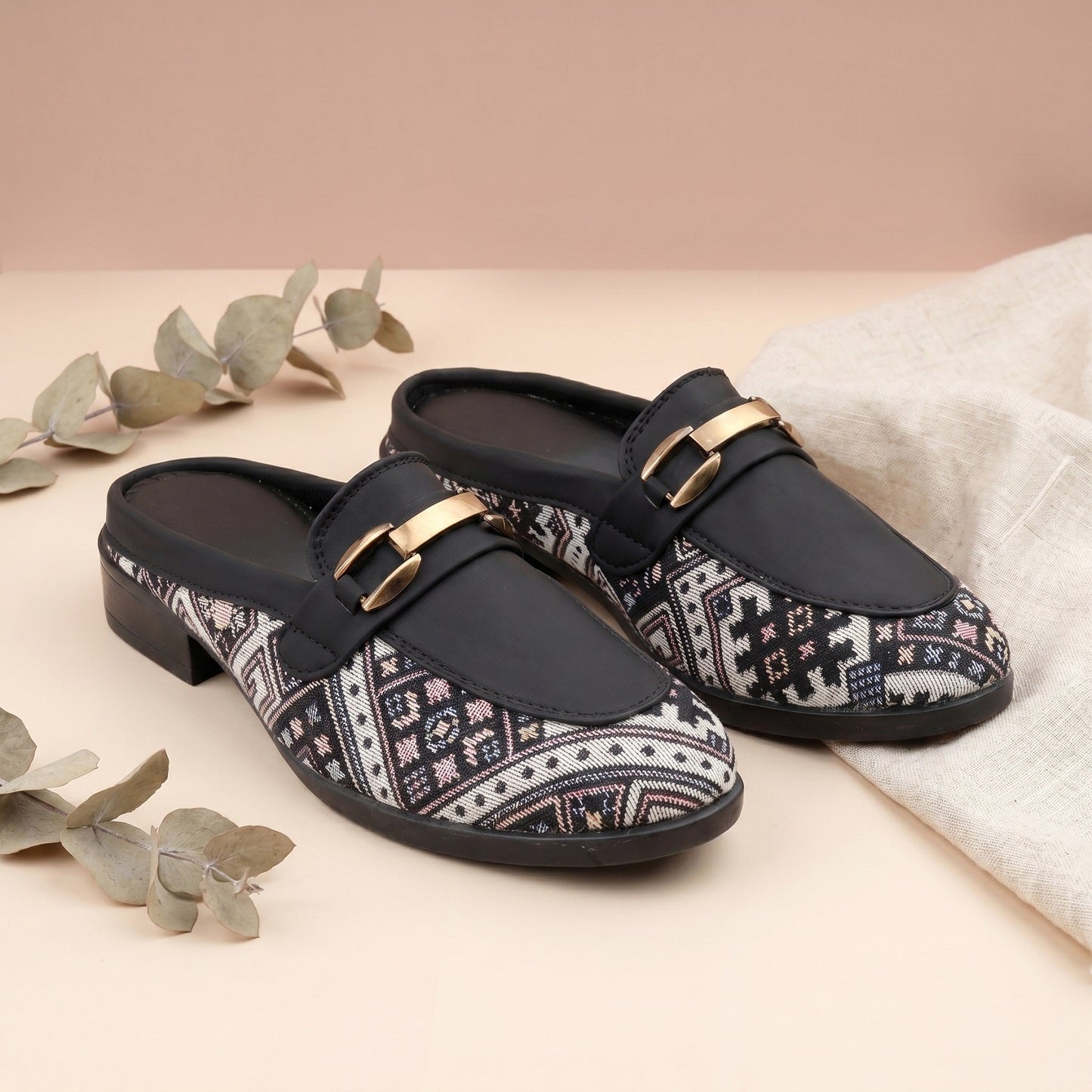 Black loafers with patterned soles and gold buckles on a beige surface with eucalyptus leaves.