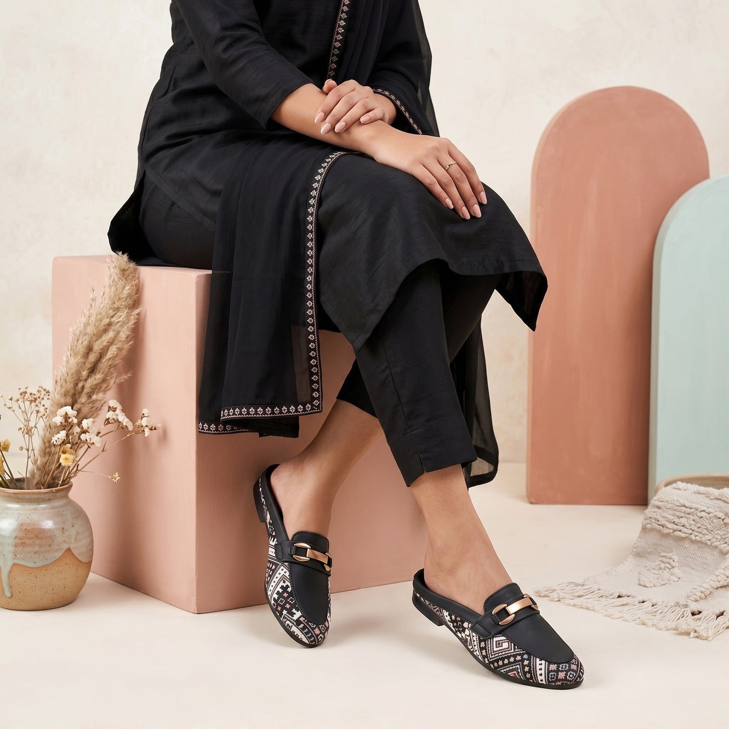 Person wearing black shoes with decorative patterns sitting on a pink block.