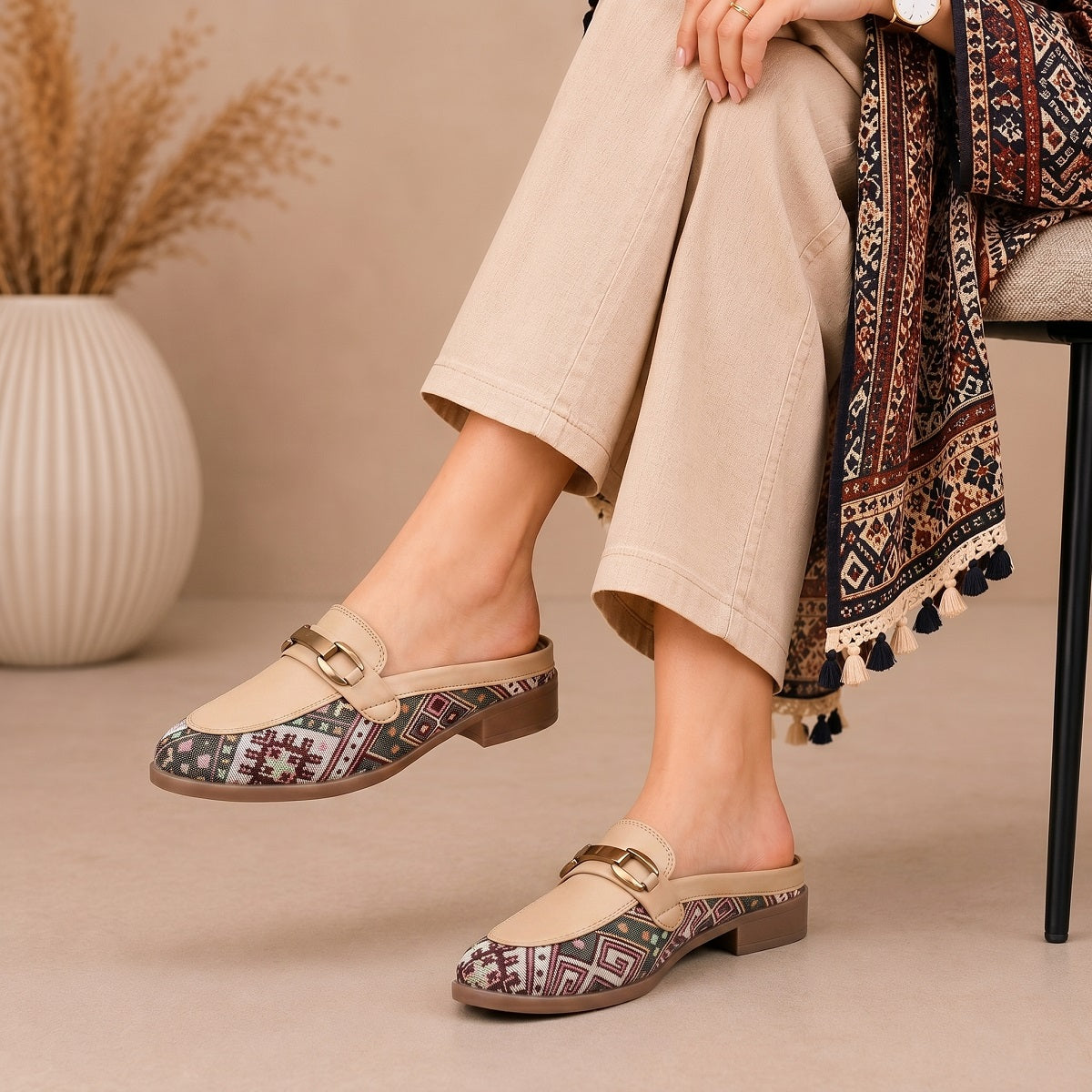 A person seated on a chair wearing beige slip-on loafers with geometric patterned toes and gold-tone buckle straps. The scene includes beige cropped pants, a patterned draped scarf, and a vase with dried grass in the background.