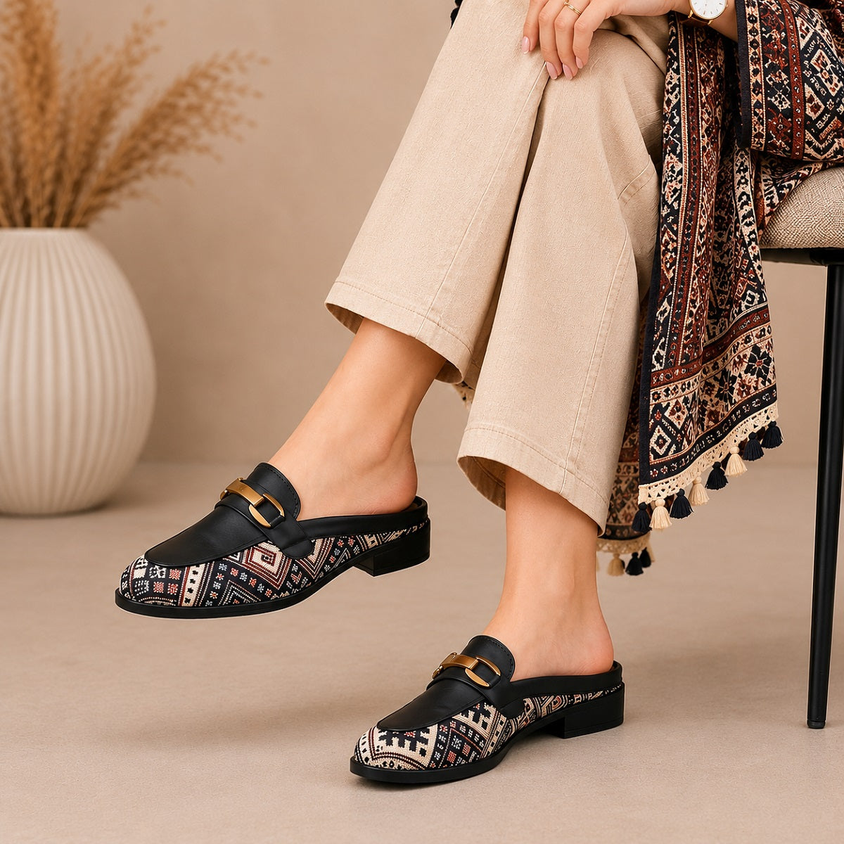 Person wearing patterned black clogs with beige pants and a decorative scarf, sitting on a chair.
