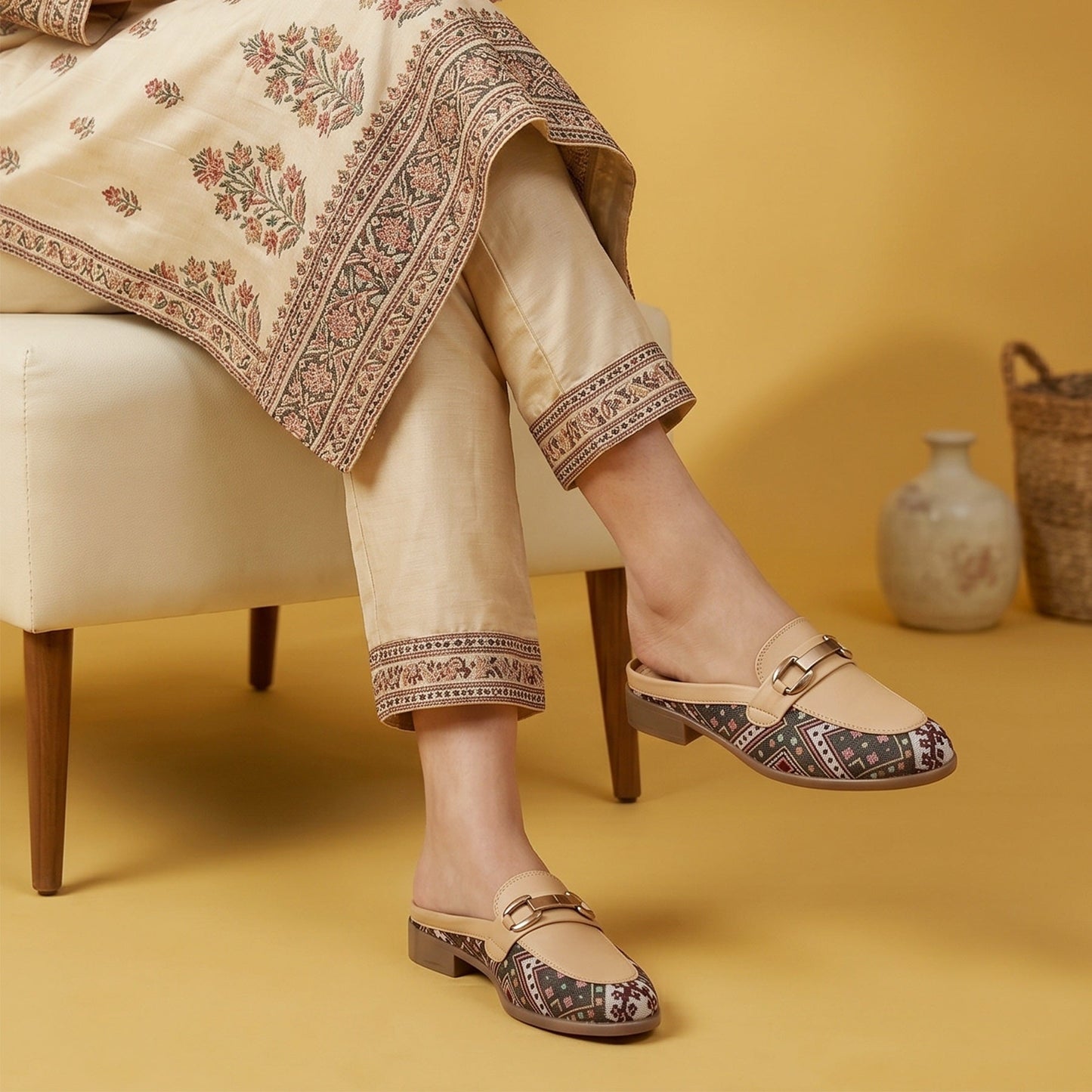 A person seated on a cream chair wearing beige embroidered trousers and matching draped fabric, paired with beige flat mule loafers that have patterned insoles and a small gold buckle detail. The scene is set against a warm mustard-yellow background with a small vase and basket.