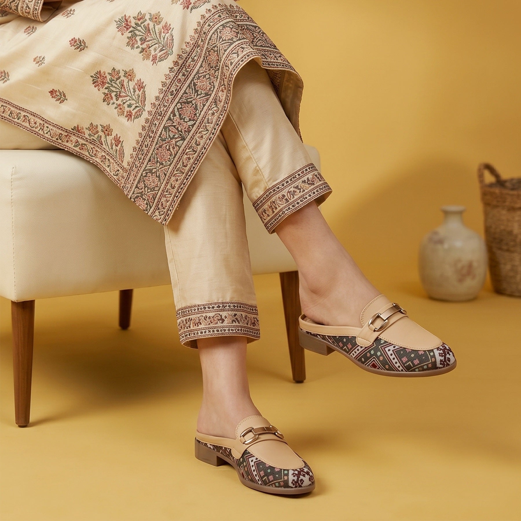 A person seated on a cream chair wearing beige embroidered trousers and matching draped fabric, paired with beige flat mule loafers that have patterned insoles and a small gold buckle detail. The scene is set against a warm mustard-yellow background with a small vase and basket.