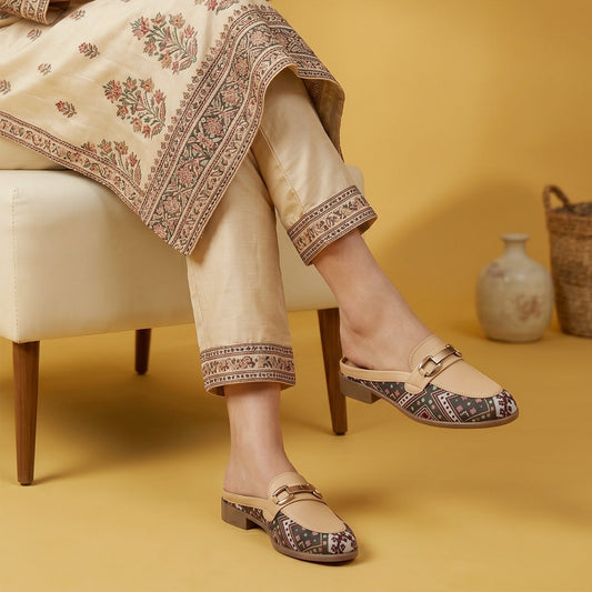 A person seated on a cream chair wearing beige embroidered trousers and matching draped fabric, paired with beige flat mule loafers that have patterned insoles and a small gold buckle detail. The scene is set against a warm mustard-yellow background with a small vase and basket.