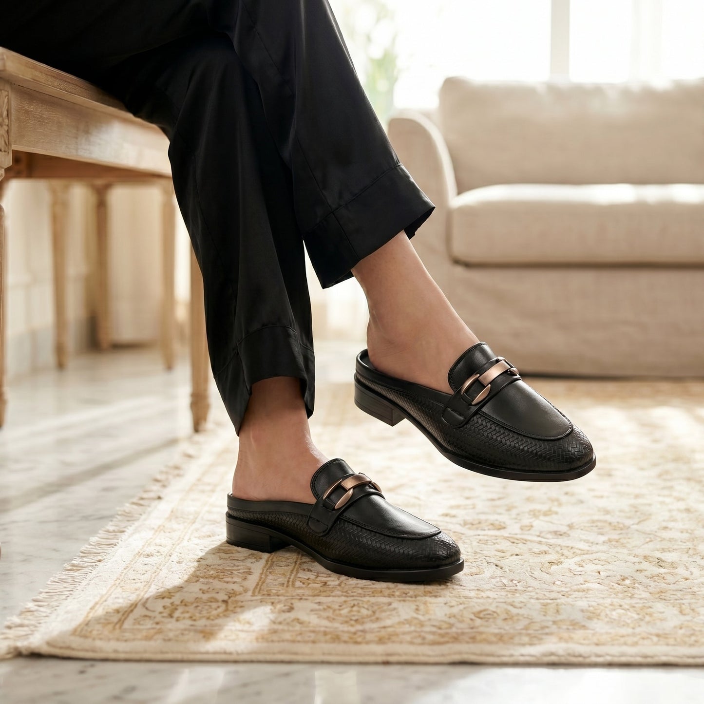 A person wearing black leather loafers with a metallic gold-tone bar detail, sitting on a wooden bench in a bright living room.