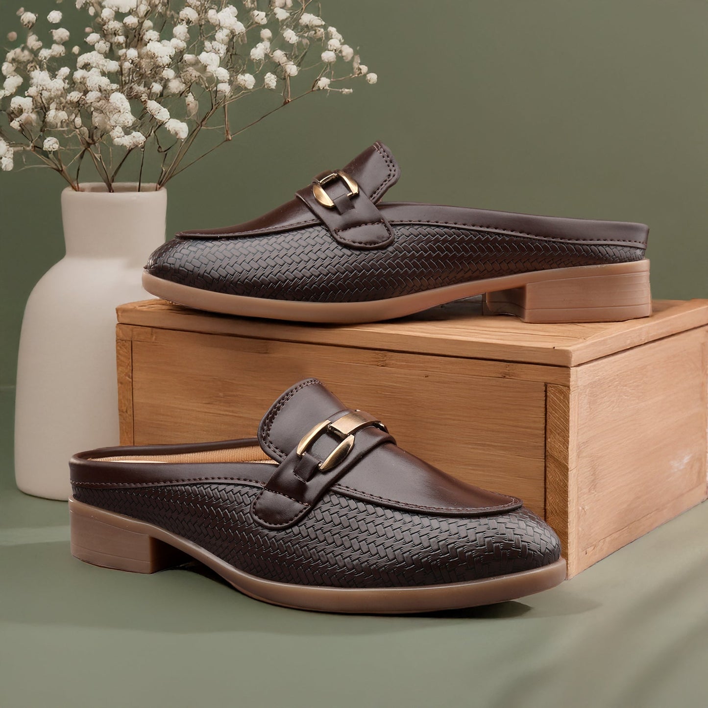 Brown woven loafers with gold buckles on a wooden box with a vase of white flowers in the background.